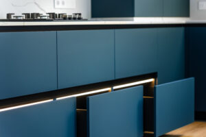 Matte blue base cabinets with a continuous LED strip under the countertop, open drawers guide the eye toward the gas cook top. SSUCv3H4sIAAAAAAAAA41PQY6DMAz8i88gBciWDV+p9hCIS60CqRKz1Qrl73UoVa+b04wnnhlvQA46cMbUqtWqbBtzKqvqokpjdVuqy6nHvnaVqREKcJYRuqppjKlqrfW3+jJKXgG9jTRAtwFN0xo5WCa/QCdKwMVh2CE6Yh/ITsJSAZEtrxGjrAkbxHsUdedvv/OW51JQwuPaZ5CKf85+CrAjLsNf9k+5yIR2jzuLdHswhvnI9nzNFTO8BxpoGY9OcoqfX3jxnHf3BPbDjdyx/DrjSjINOQvW++StQ3fIaz8T85sG/CV8fH6nlJ6DMFTshAEAAA==